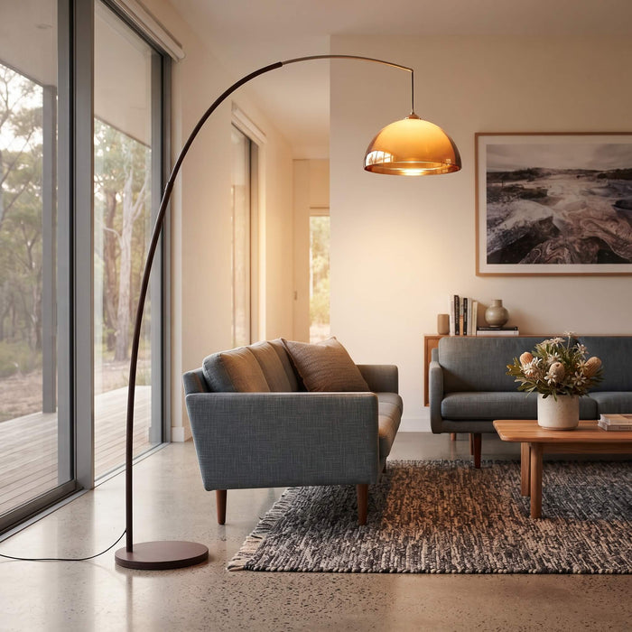 A modern curved floor lamp with a warm glowing dome shade stands in a contemporary living room next to a blue-gray sofa and wooden coffee table.