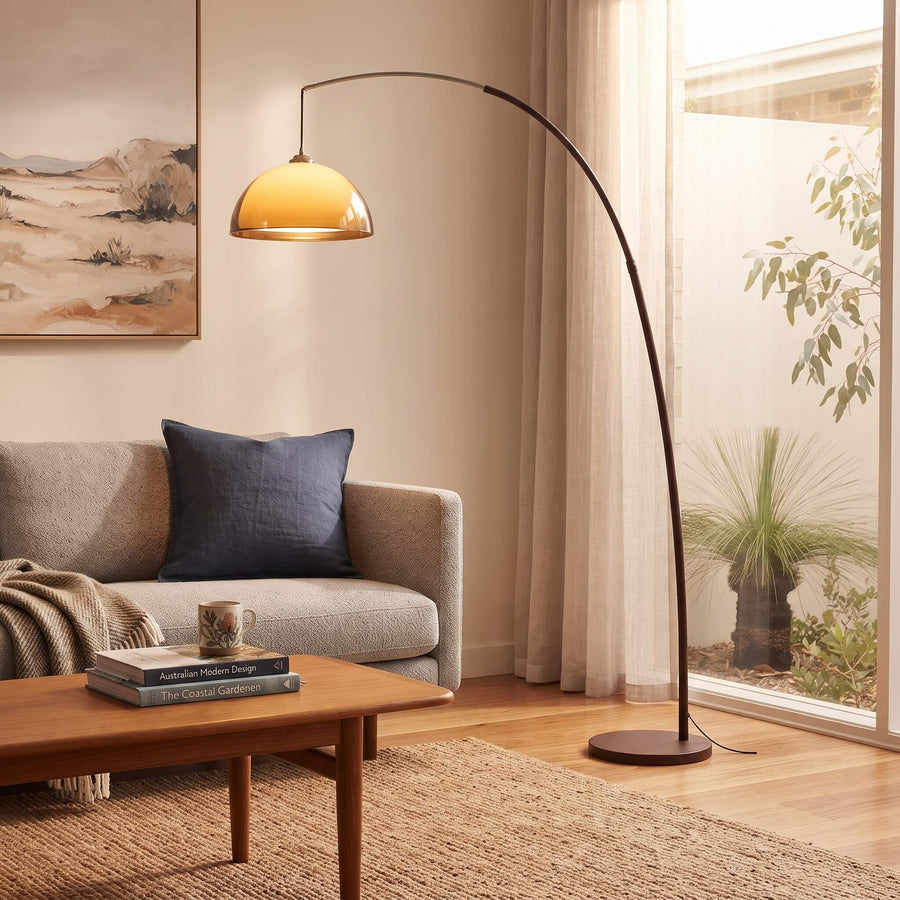 An arched floor lamp with a warm glowing dome shade stands in a modern living room next to a window with sheer curtains, with a sofa and coffee table in the background.