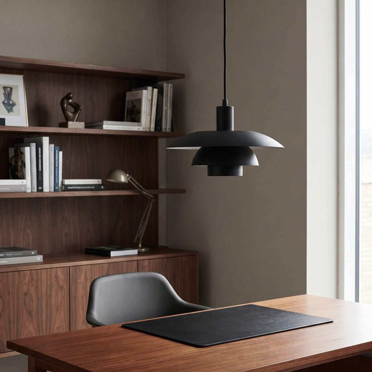 A modern black pendant light with a layered, petal-like shade hangs over a wooden desk in a minimalist home office setting with a dark wood bookshelf and chair.