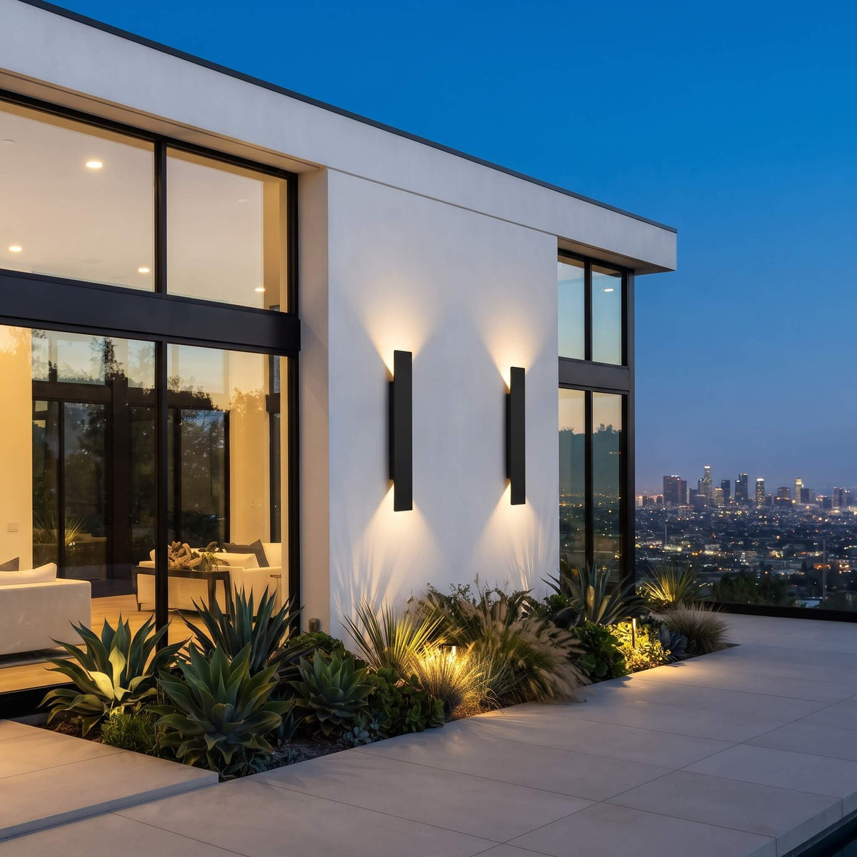 Modern black rectangular wall sconces mounted on a white stucco exterior wall of a contemporary house, casting warm light onto the wall and plants at dusk, with a city skyline visible in the background.
