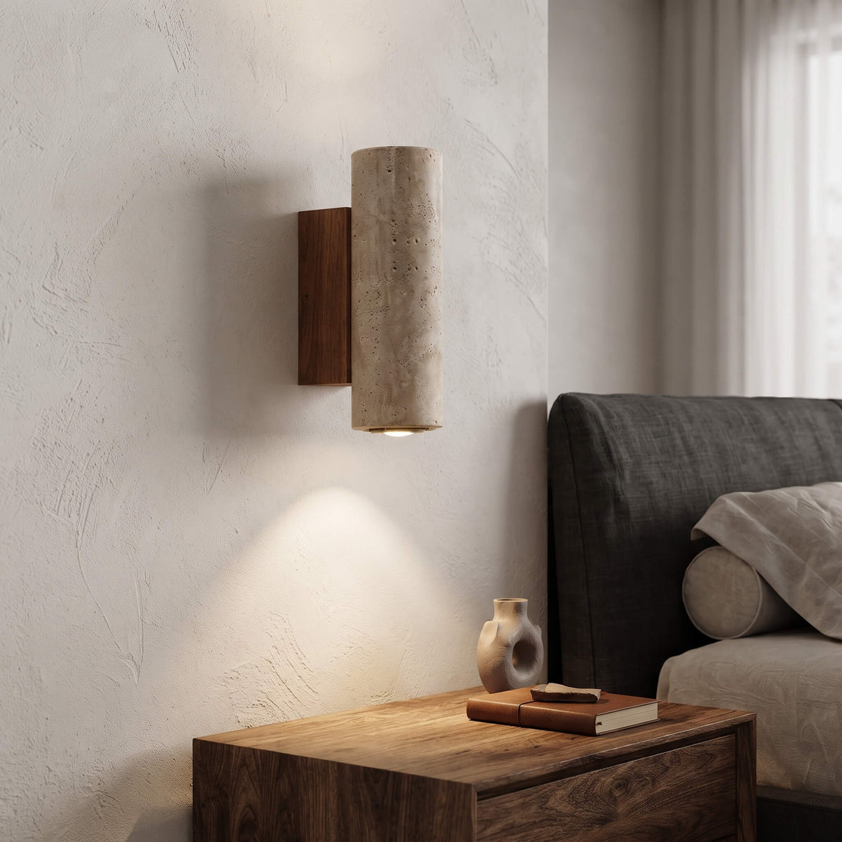 A modern cylindrical wall sconce with a textured beige stone or concrete shade and a dark brown wooden backplate, mounted on a textured off-white wall above a wooden bedside table.