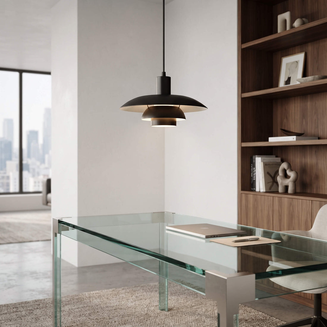 A modern black pendant light with a layered, saucer-like shade hangs over a glass-top desk in a bright, contemporary home office. The room features a white wall, a large window with a city skyline view, and a tall wooden bookshelf on the right.