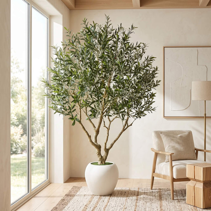 A tall artificial olive tree with green leaves and brown branches, potted in a white ceramic pot, placed in a bright, modern room next to a large window.