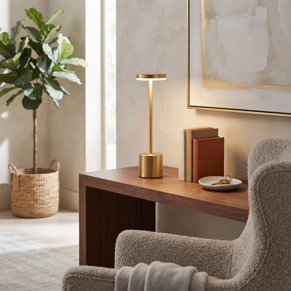 A modern table lamp with a brushed gold cylindrical base and a slim matching stem lamp head, lit to cast a warm glow, sitting on a wooden desk in a styled home office setting.