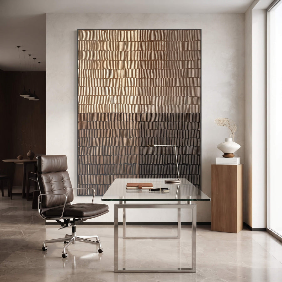 A modern home office interior featuring a large vertical wall art piece with a textured vertical stripe pattern in warm beige, brown, and charcoal tones, positioned behind a glass-top desk and a black leather office chair.