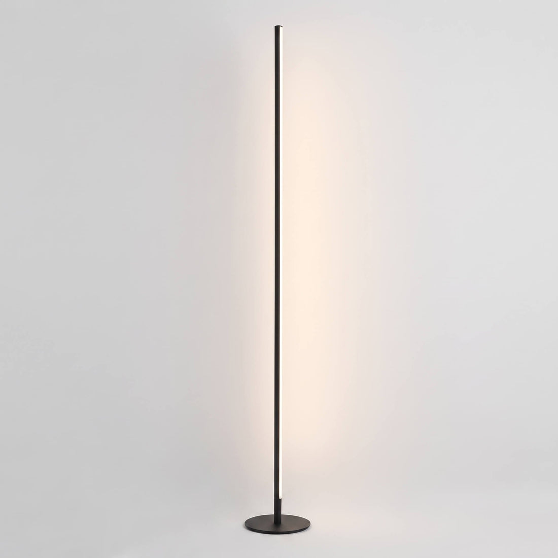 A modern slim floor lamp with a dark metal circular base and a long vertical rod emitting a warm light, shown against a plain light gray background.