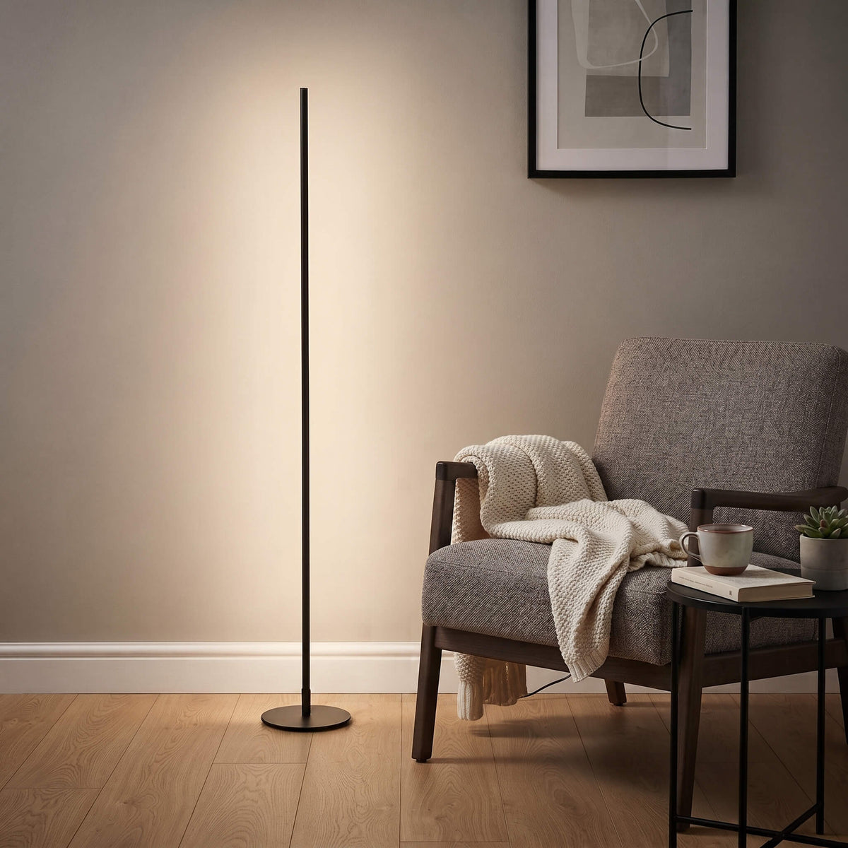 A tall, slim black floor lamp stands on a light wood floor next to a gray upholstered armchair with a cream throw blanket, in a styled living room setting.