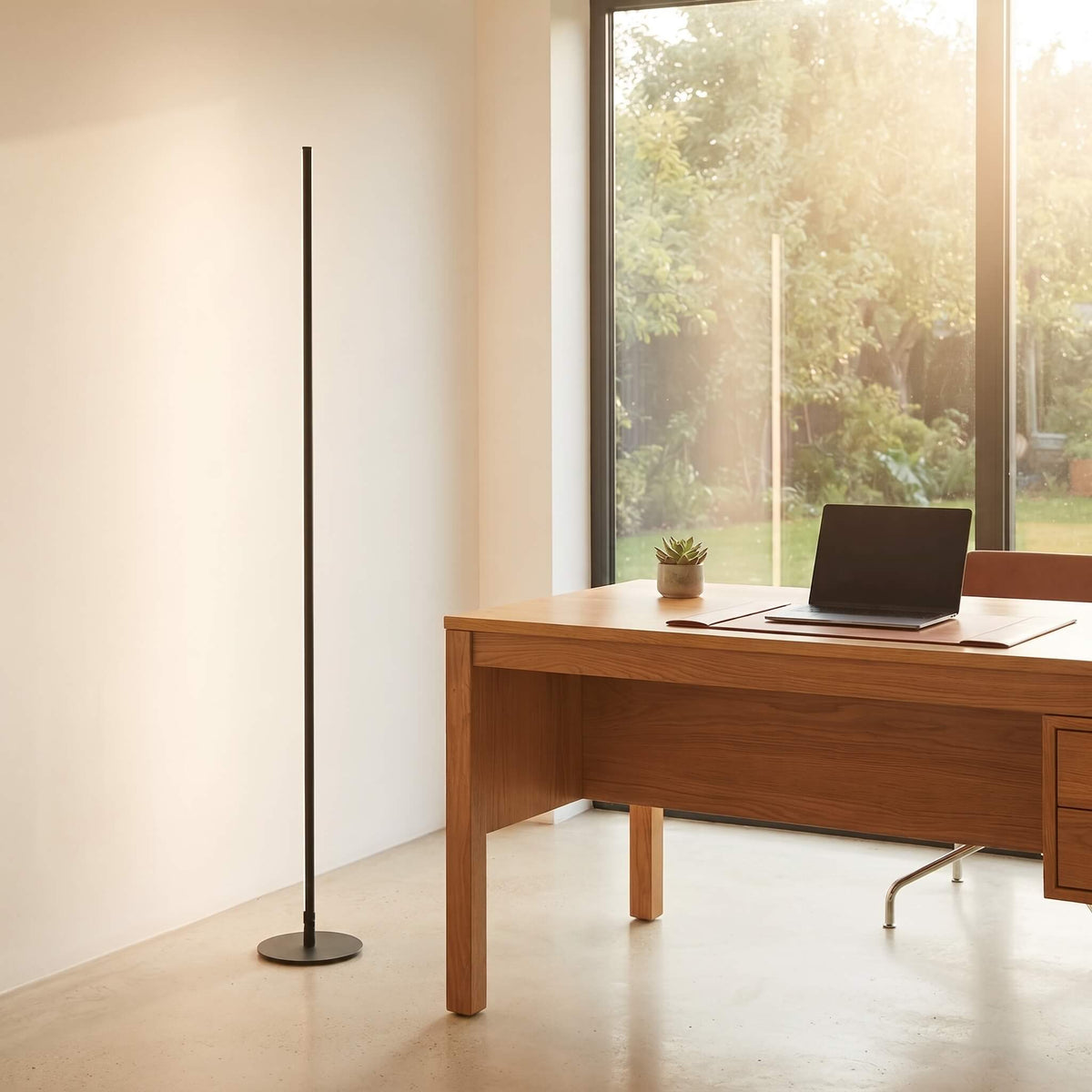 A minimalist black floor lamp stands on a light concrete floor next to a wooden desk in a bright, modern room with large windows showing a garden view.