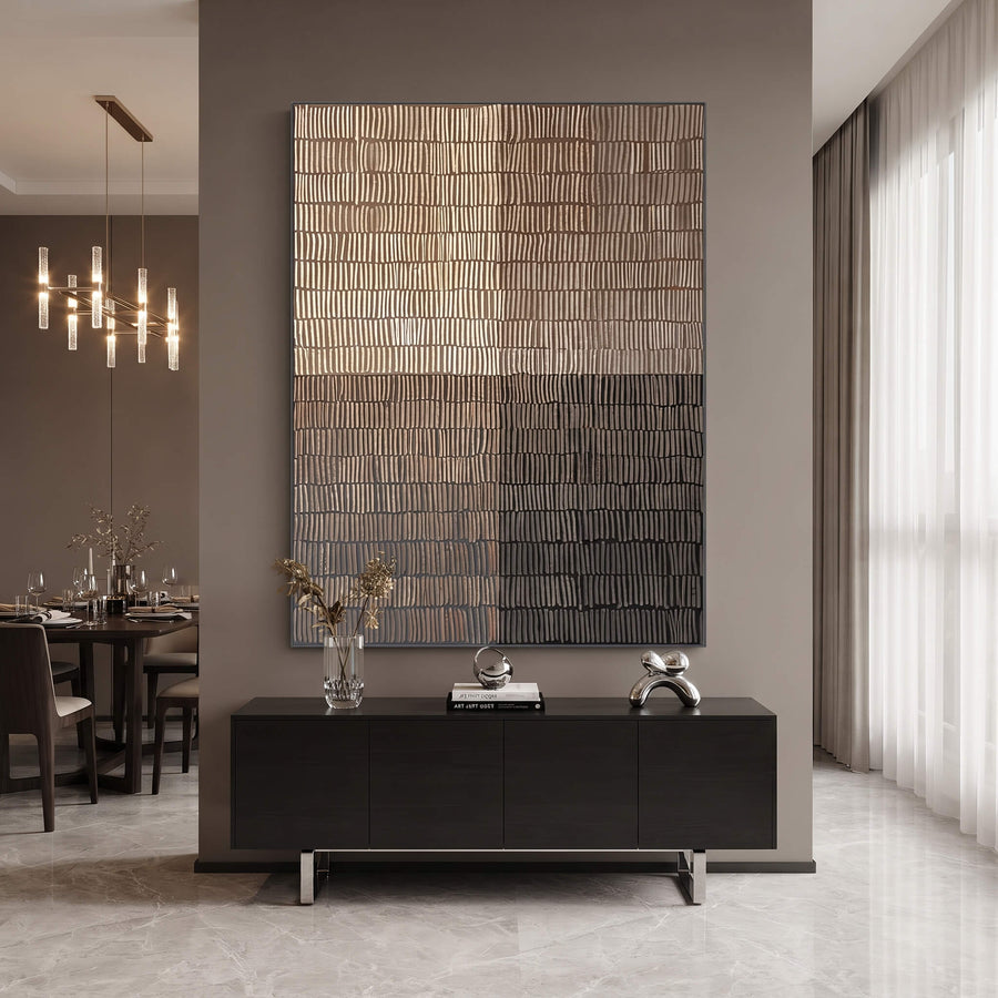A large rectangular wall art piece with a textured, ribbed metallic surface in warm gold, bronze, and dark gray tones, displayed above a black console table in a modern dining room setting.