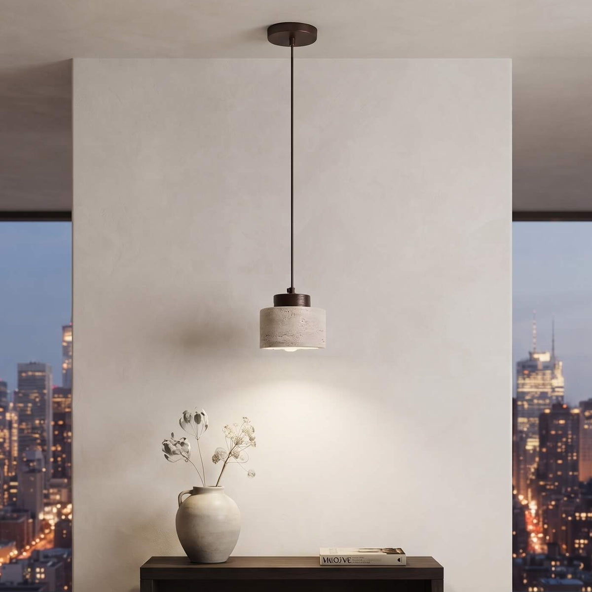 A modern cylindrical pendant light with a dark metal rod and a textured light gray shade hangs over a dark wood console table in a minimalist room. Large windows in the background show a city skyline at dusk.