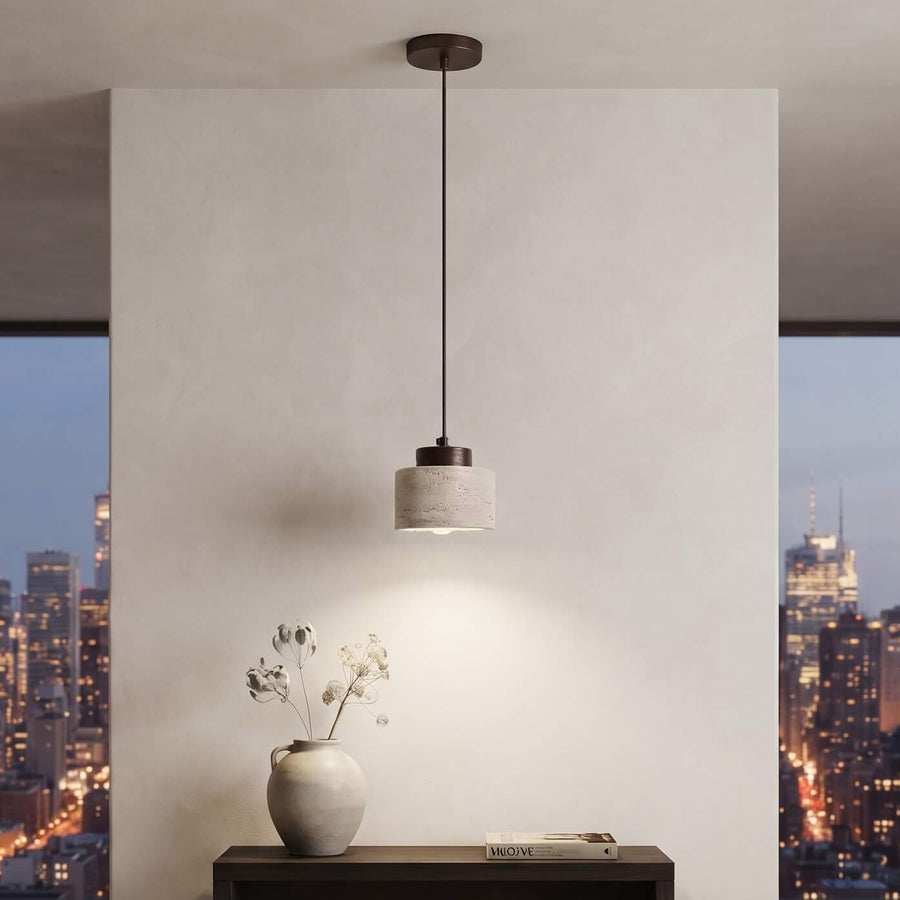 A modern cylindrical pendant light with a dark metal rod and a textured light gray shade hangs over a dark wood console table in a minimalist room. Large windows in the background show a city skyline at dusk.