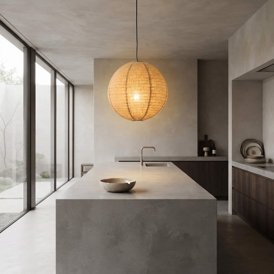 A spherical woven pendant light glowing with warm light, hanging in a modern minimalist concrete kitchen with a central island and large windows.