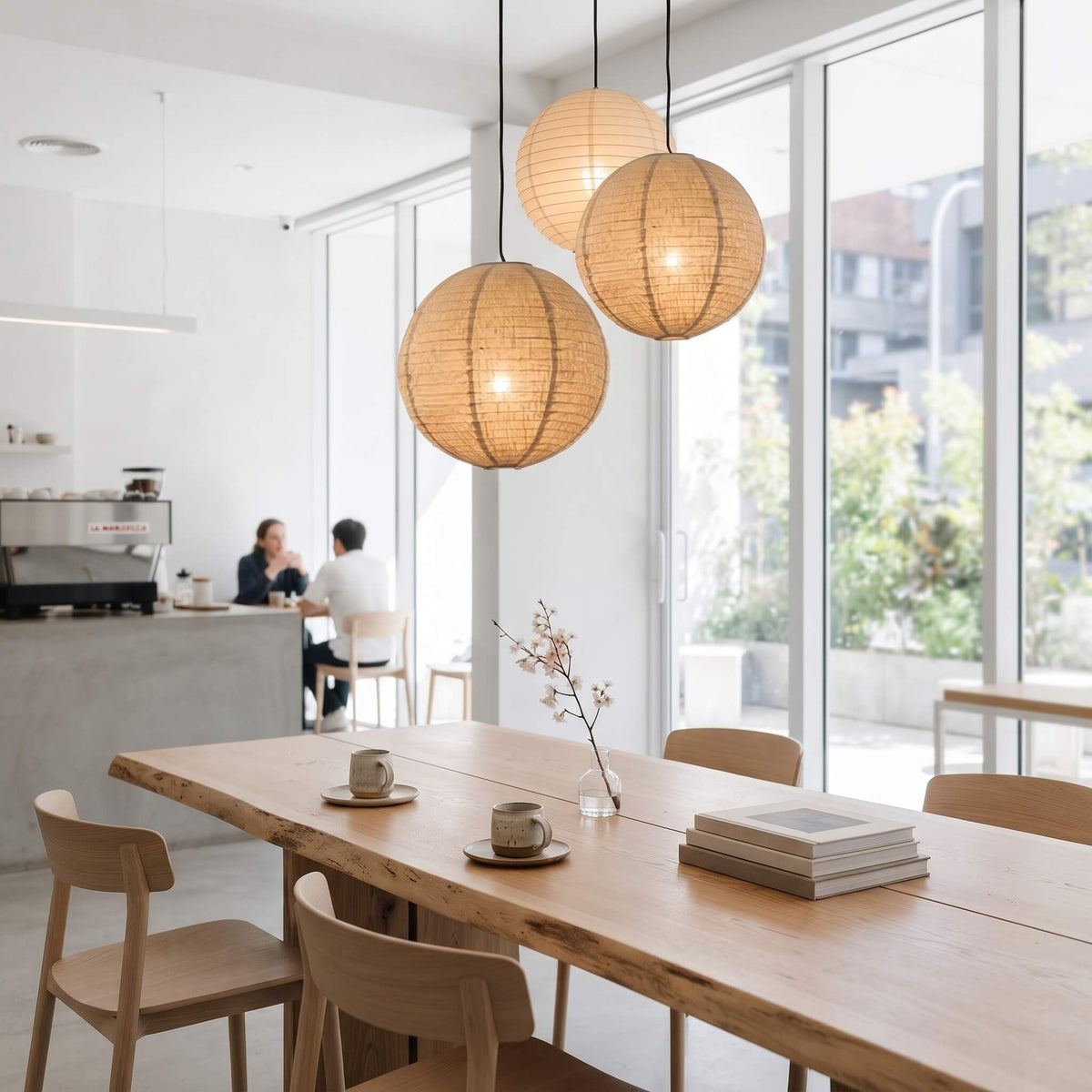 Three spherical woven rattan pendant lights hang over a light wood dining table in a bright, modern cafe interior with large windows and a counter in the background.