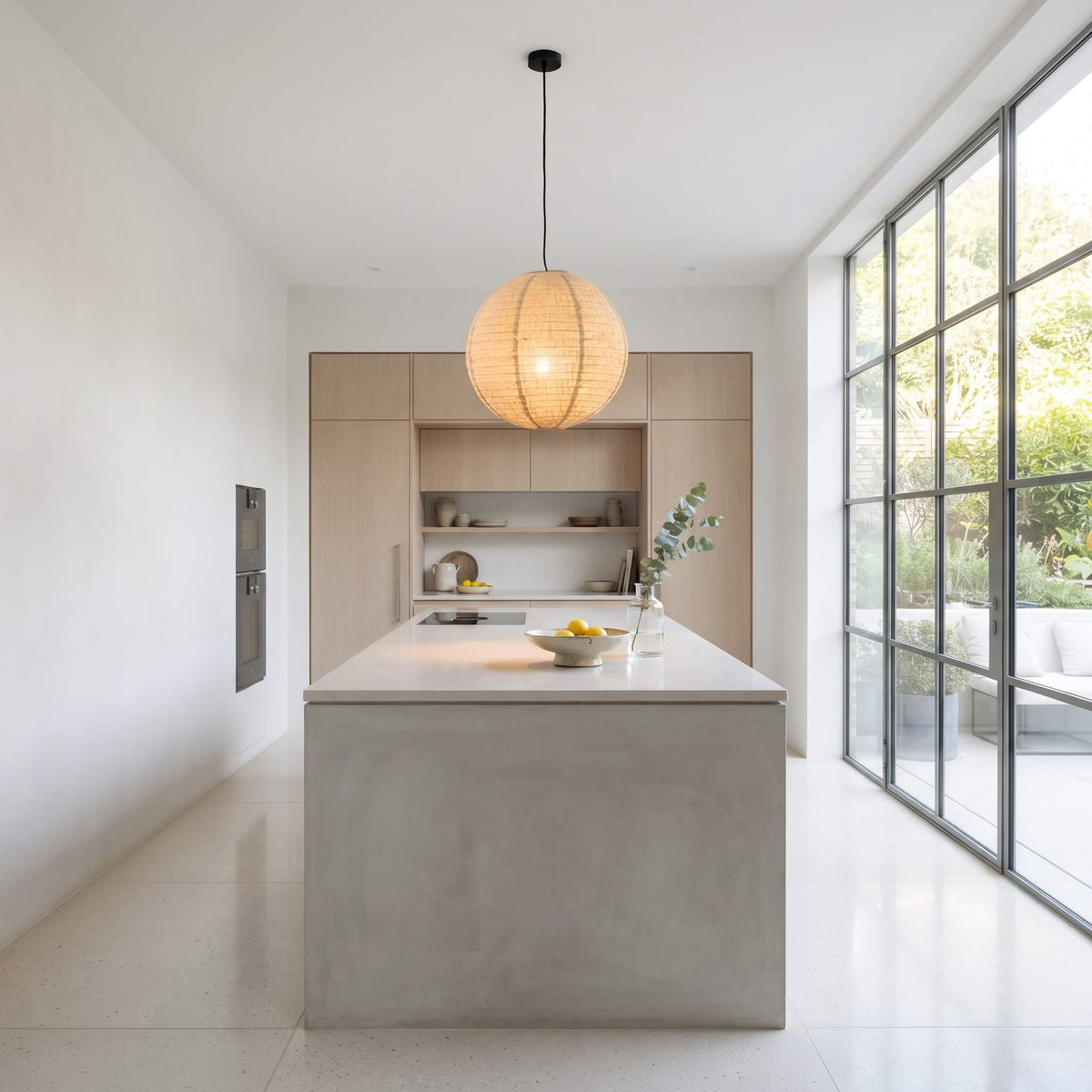 A modern kitchen island with a light gray concrete-look base and a white countertop, featuring a large woven pendant light hanging above. The background shows light wood cabinetry, a built-in sink, and a large grid-style window looking out to greenery.