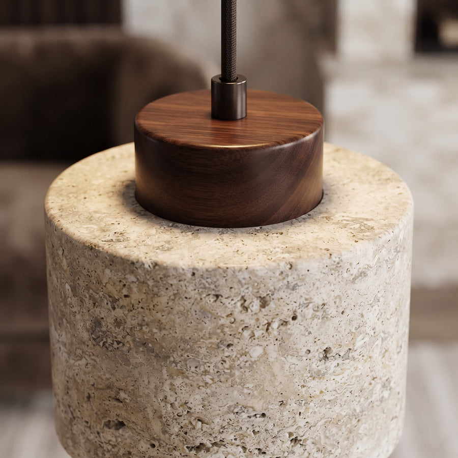 A close-up of a cylindrical pendant light with a textured beige stone base and a dark brown wooden top, with a dark metal cord extending upward against a softly blurred indoor background.