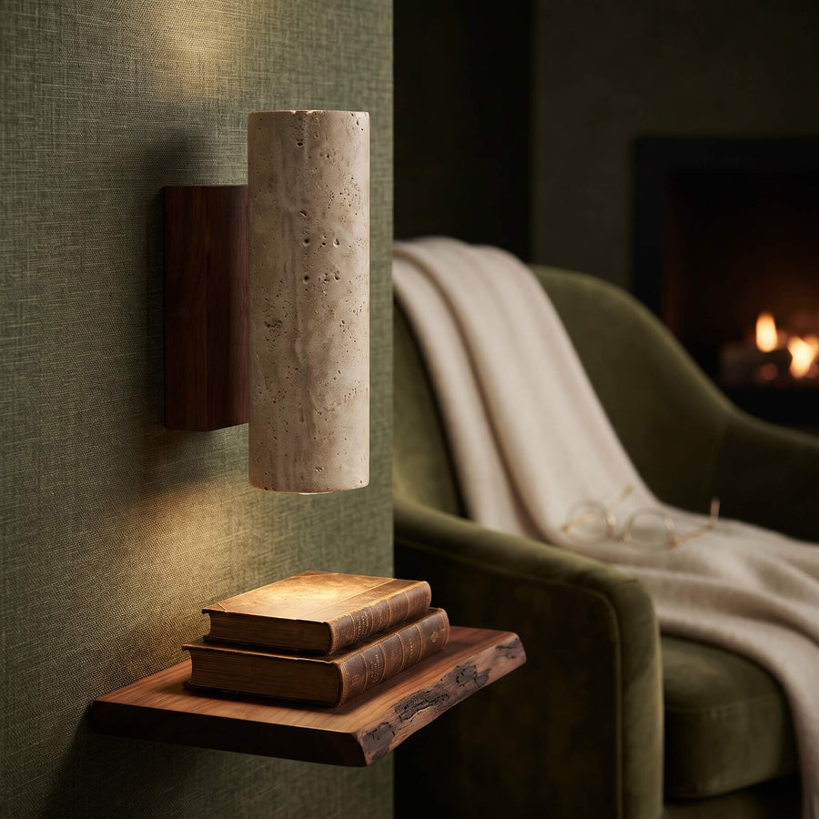 A modern wall-mounted sconce with a cylindrical textured stone shade and a dark wood backplate, shown lit in a cozy living room setting next to a green armchair and a floating wooden shelf with books.