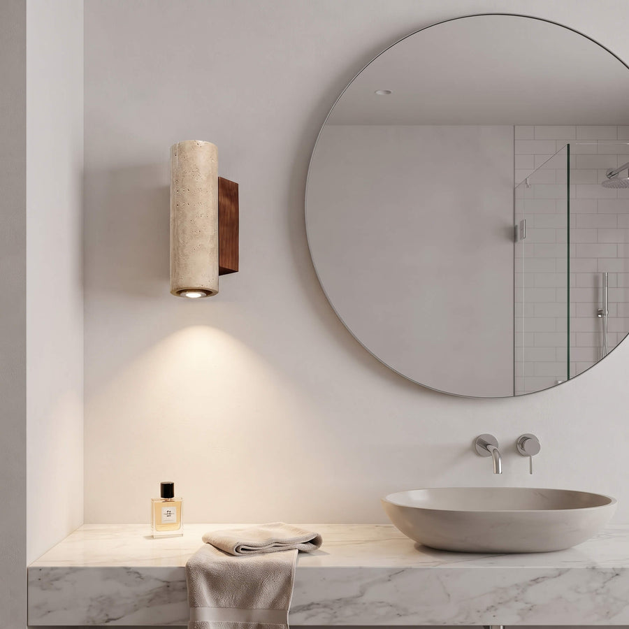 A modern cylindrical wall sconce with a textured beige stone-like finish and a dark wood accent, mounted on a bathroom wall above a marble vanity with a vessel sink and round mirror.