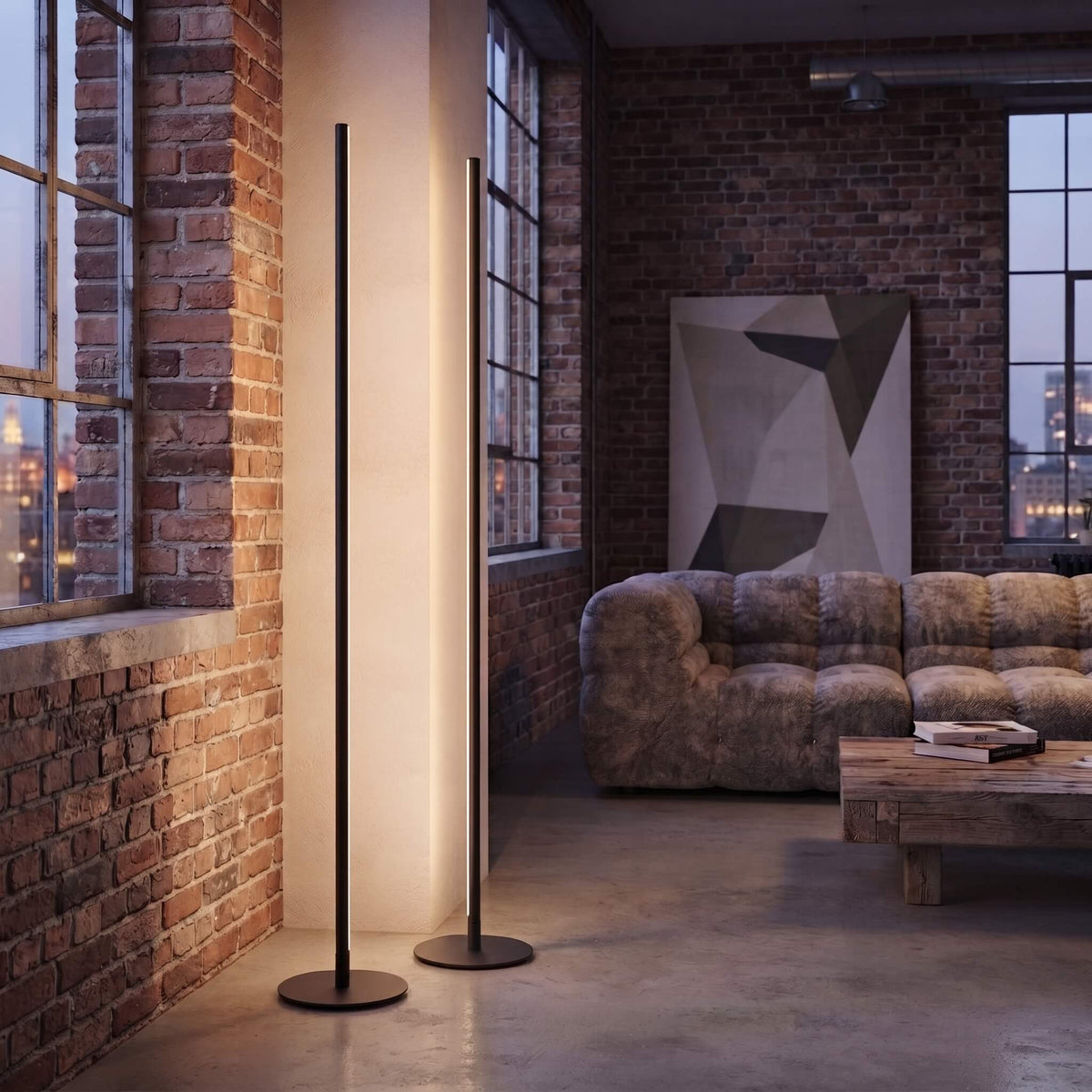 A modern minimalist black floor lamp with a slim pole and circular base stands in a loft-style living room with brick walls, a textured sofa, and a coffee table.