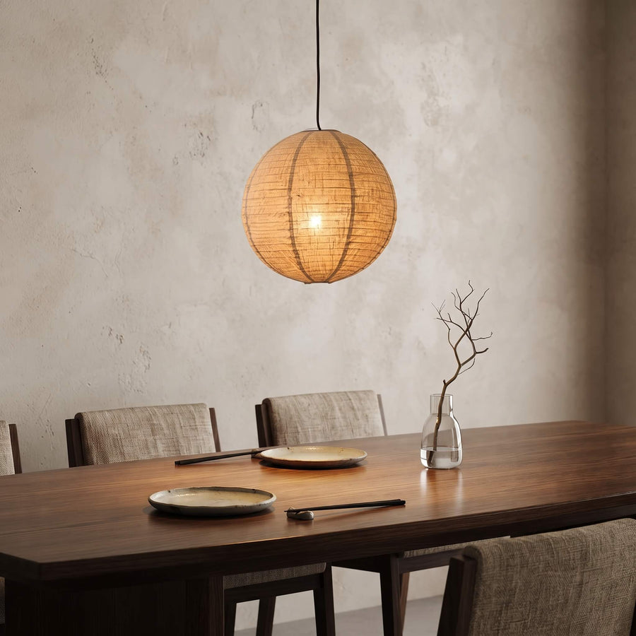 A spherical woven pendant light in a warm natural straw color hangs over a dark wood dining table with chairs, plates, and a small glass vase in a softly lit interior.