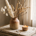 Vase with dried flowers and a lit candle on a wooden table.