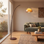 An arched floor lamp with a dark curved metal stand and a warm amber dome shade stands in a modern living room beside large windows, casting light over a green sofa and wooden coffee table.