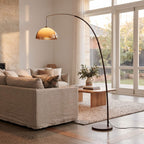 A modern floor lamp with a tall black arched stand and a warm amber dome shade stands in a bright, contemporary living room next to a beige sofa and wooden coffee table.