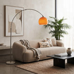A modern living room scene featuring a beige boucle sofa with a gray throw blanket, a black coffee table, a potted palm, and a tall floor lamp with a glowing orange shade.