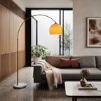 A modern floor lamp with a curved silver metal arm and a glowing orange dome shade stands in a contemporary living room next to a gray sofa with a beige throw blanket.