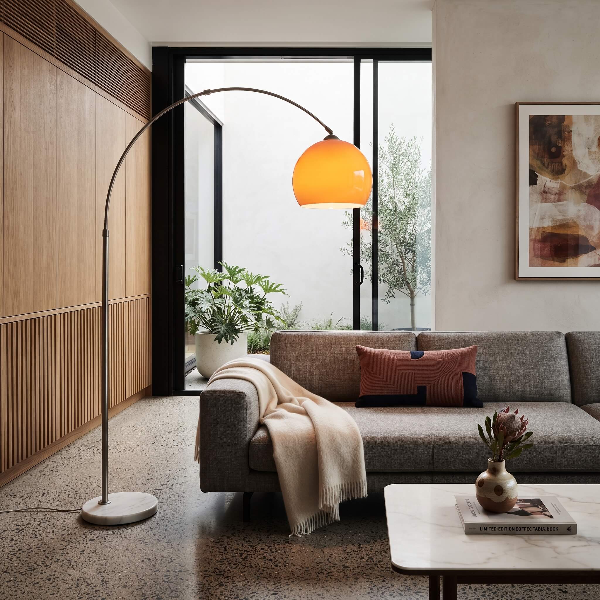 A modern floor lamp with a curved silver metal arm and a glowing orange dome shade stands in a contemporary living room next to a gray sofa with a beige throw blanket.