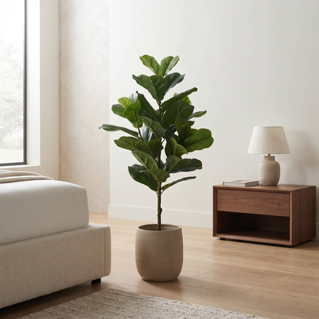 A tall fiddle-leaf fig plant with large green leaves in a beige ceramic pot, placed in a modern bedroom next to a wooden nightstand and lamp.