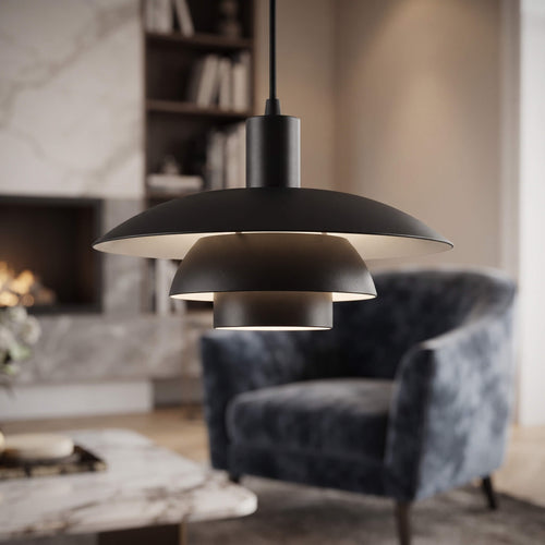 A modern black pendant light with a wide, layered disc design hanging in a cozy living room with a blue-gray armchair and fireplace in the background.