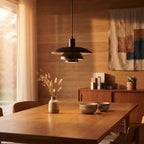 A modern black pendant light with a wide, tiered shade hangs over a wooden dining table in a warm, sunlit room with wood-paneled walls and neutral decor.
