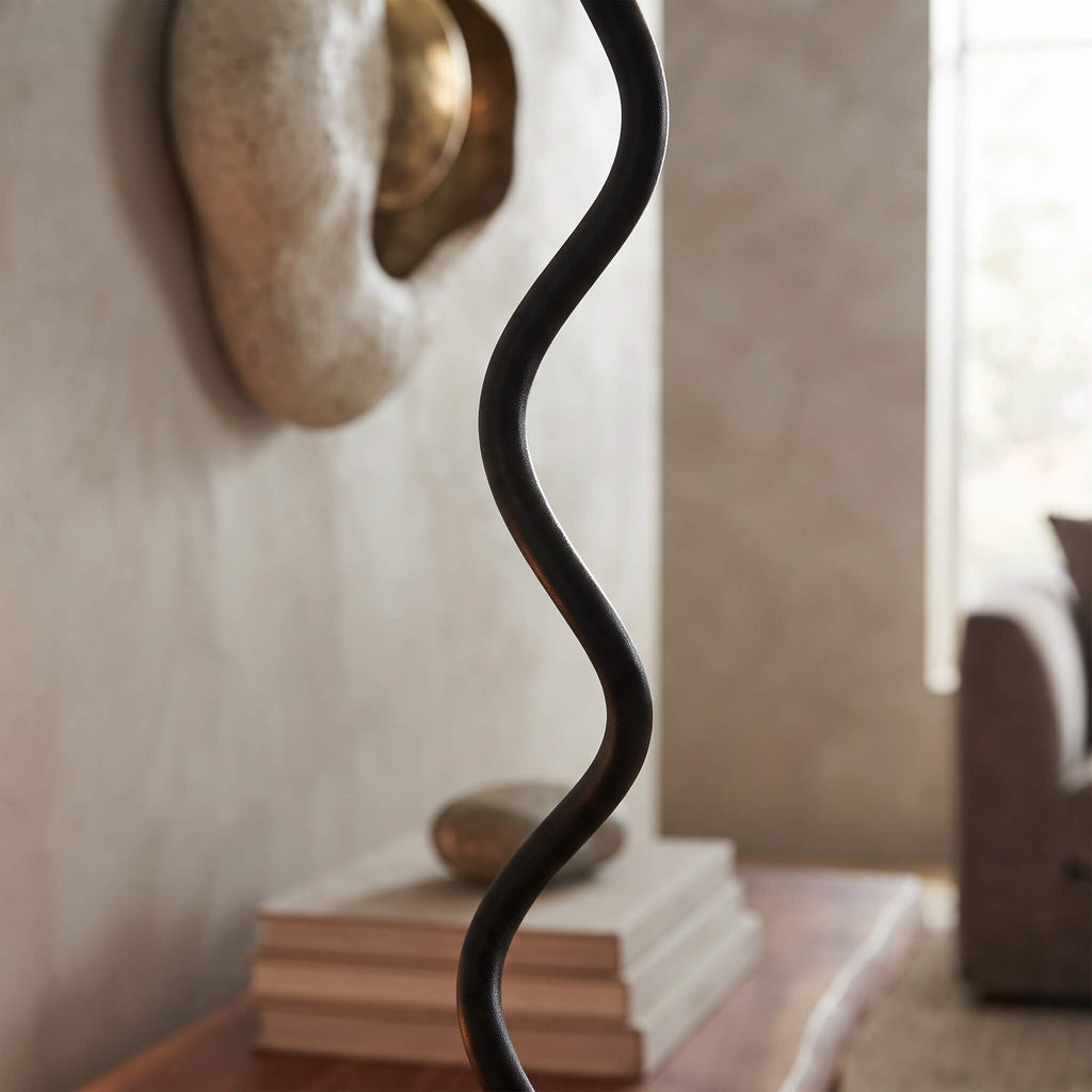 A close-up of a black metal floor lamp with a wavy, sculptural cord-like shade in a softly lit living room setting.