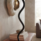 A close-up of a black metal floor lamp with a wavy, sculptural cord-like shade in a softly lit living room setting.