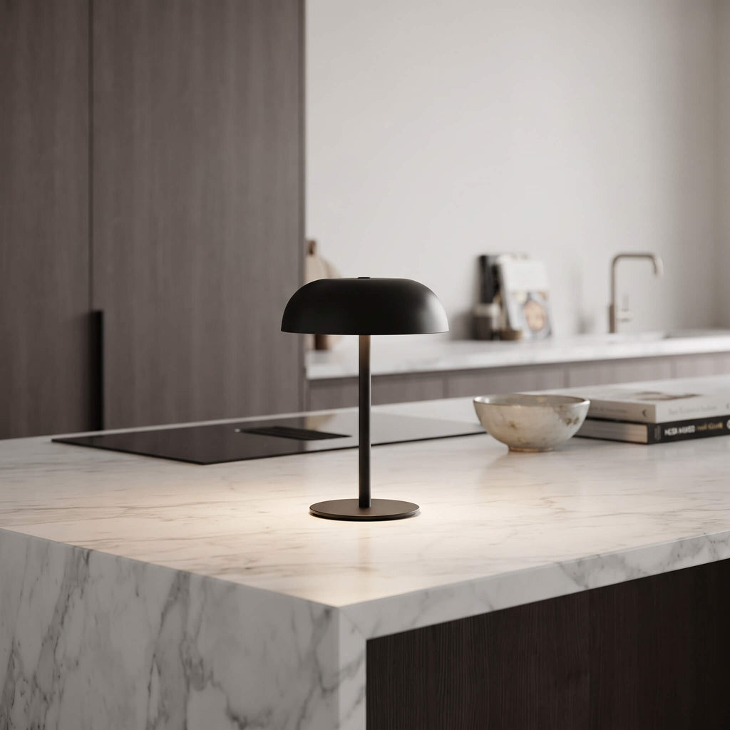A modern black table lamp with a dome-shaped shade and slim base sits on a white marble kitchen island in a minimalist kitchen setting.