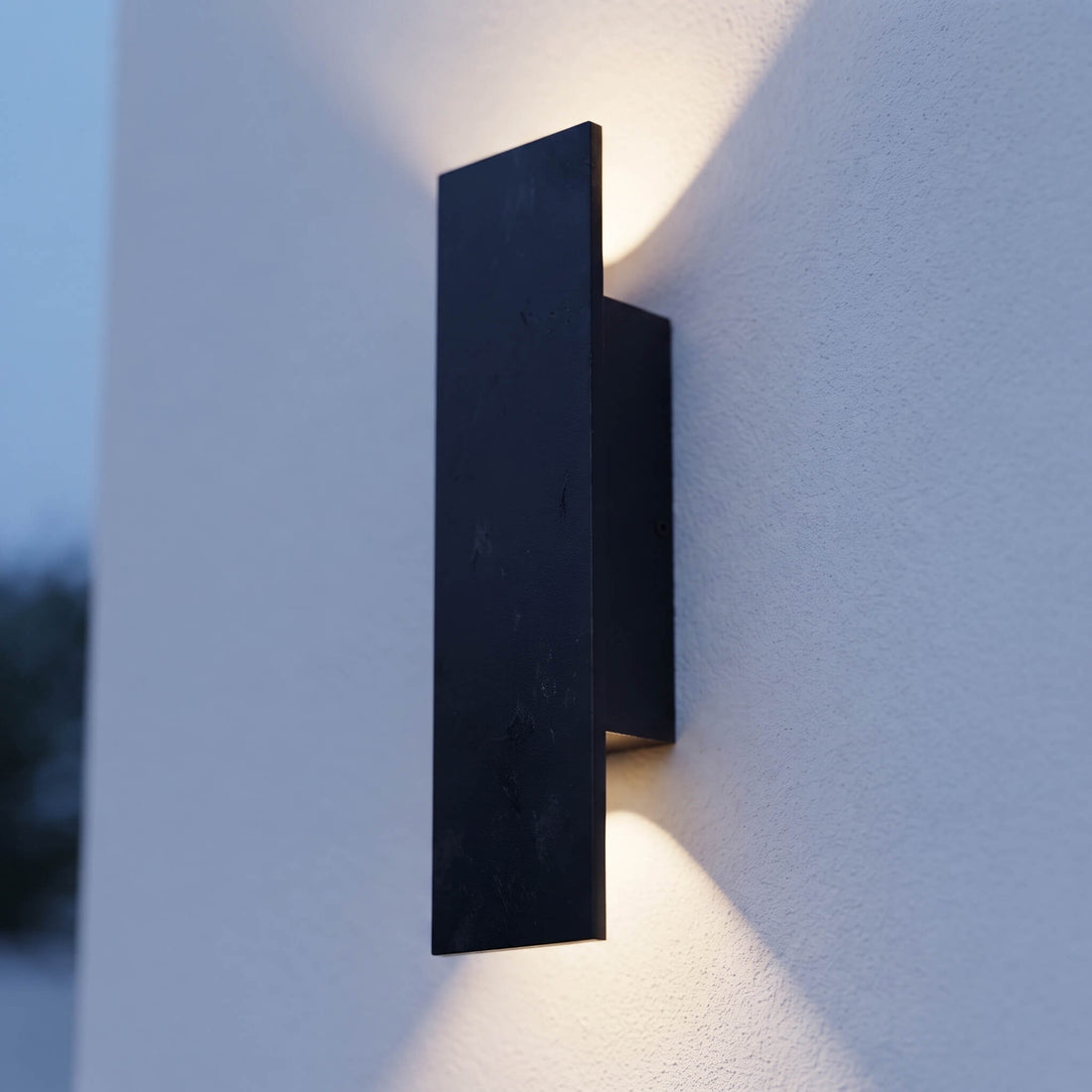 A modern matte black rectangular wall sconce mounted on a textured white wall, casting a warm light downward and upward.