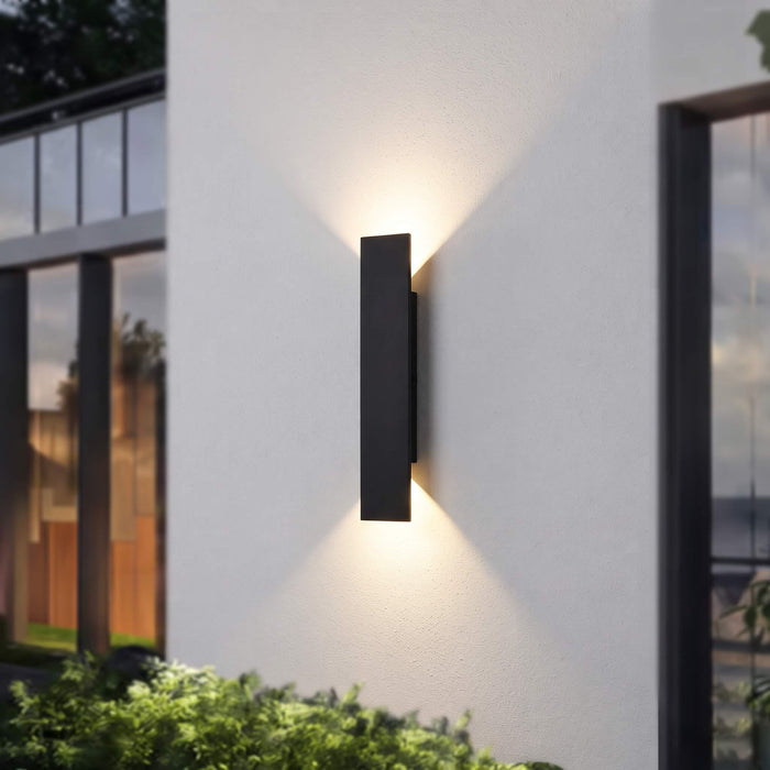 A modern vertical rectangular outdoor wall light in matte black, mounted on a white stucco exterior wall and casting a warm light downward and outward. The scene includes windows, greenery, and a softly blurred architectural background.