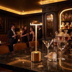 A modern minimalist table lamp with a brushed gold base and a glowing rectangular LED light bar, styled on a dark marble bar counter in a luxurious bar setting with cocktail glasses and blurred patrons in the background.