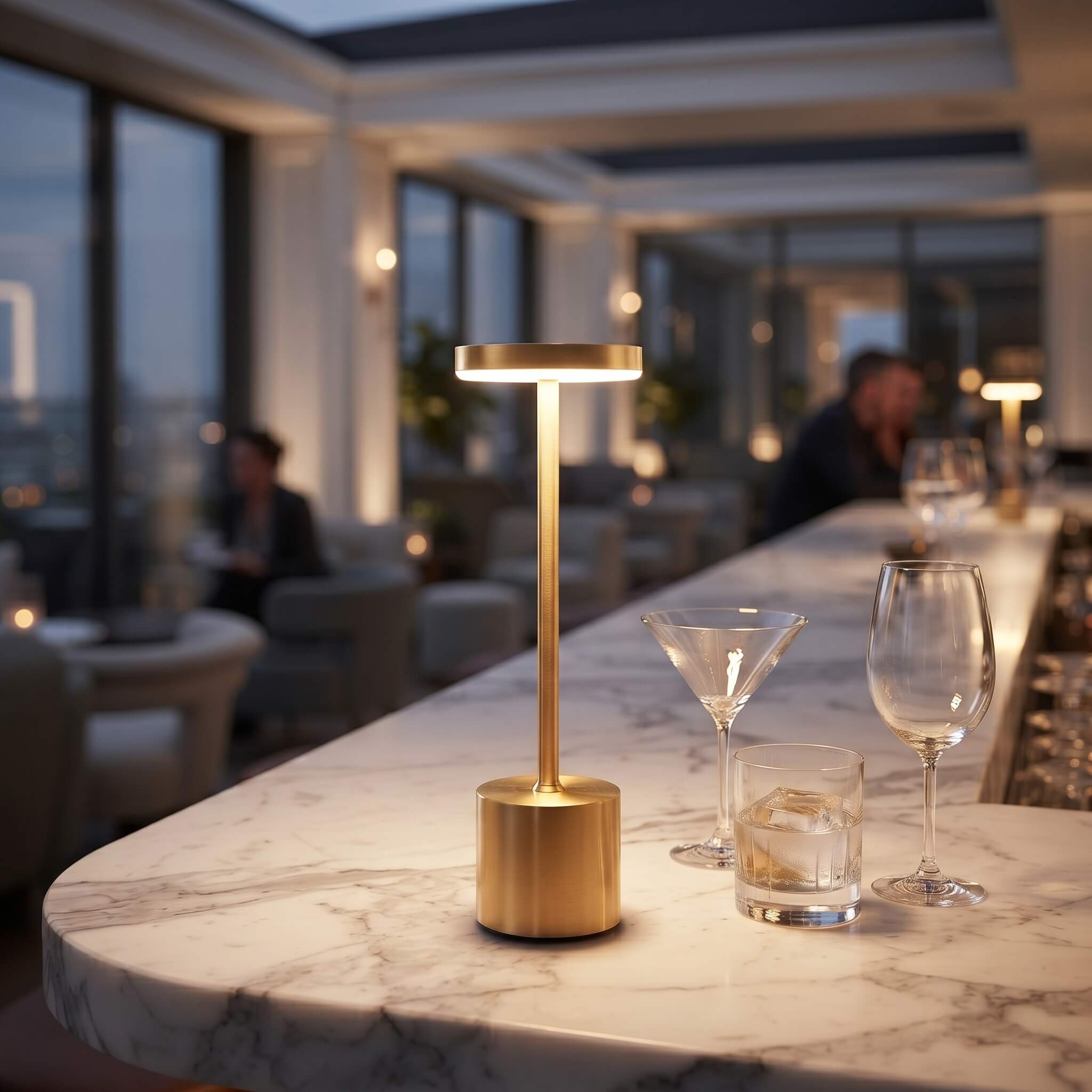 A modern gold LED table lamp with a cylindrical base and a flat rectangular light head, glowing on a white marble bar counter in a softly blurred upscale restaurant or bar setting.