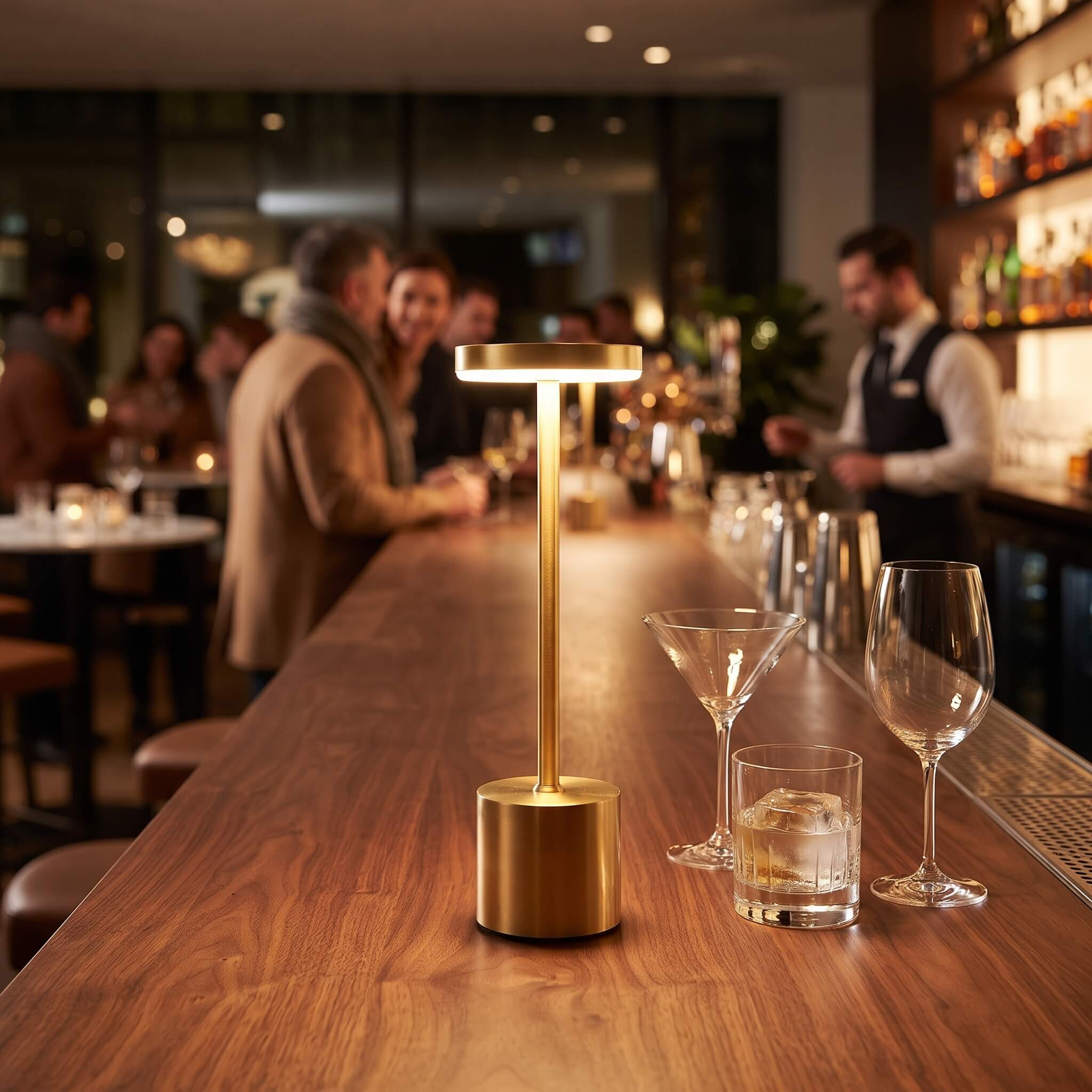 A modern gold LED table lamp with a cylindrical base and a slim rectangular light bar, placed on a wooden bar counter in a blurred bar setting with cocktail glasses nearby.