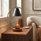 A modern table lamp with a curved brass arc and a black conical shade sits on a wooden side table in a cozy room setting next to a lit candle, books, and a cream armchair.