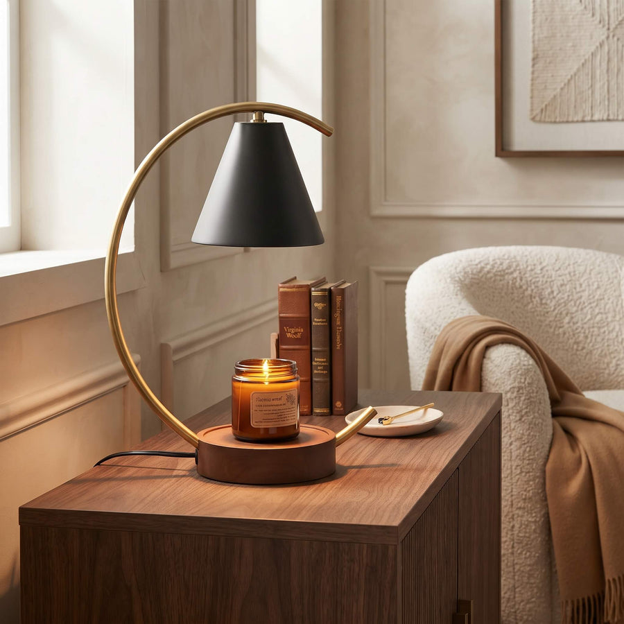 A modern table lamp with a curved brass arc and a black conical shade sits on a wooden side table in a cozy room setting next to a lit candle, books, and a cream armchair.
