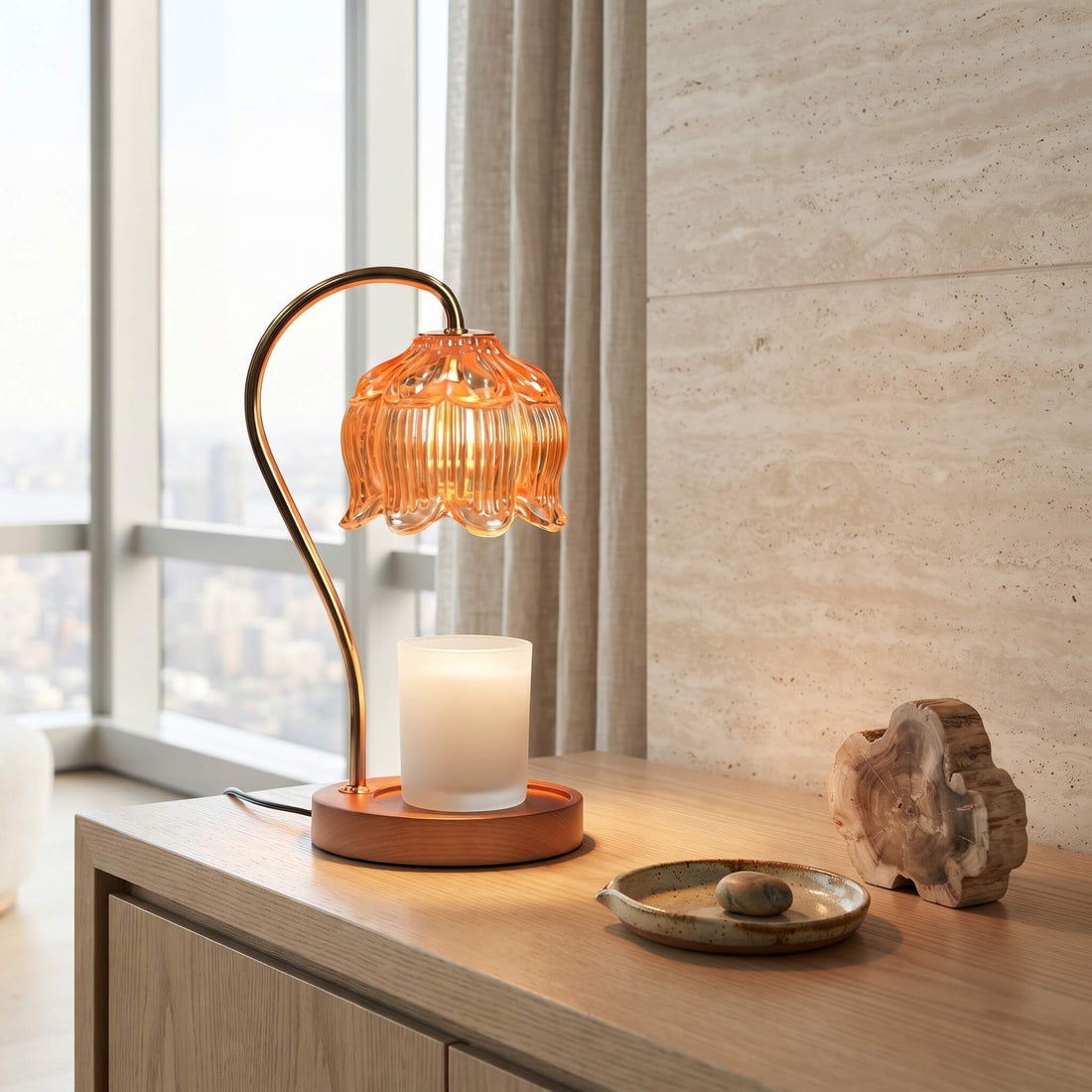 A modern table lamp with a warm orange glass flower-shaped shade and a curved copper-toned metal stem, shown lit on a wooden console table in a bright room with a window, curtain, and decorative objects.