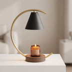 A modern floating candle table lamp with a matte black conical shade, a curved gold metal frame, and a brown wooden base holding a lit amber glass jar candle, displayed on a white table in a softly blurred interior setting.