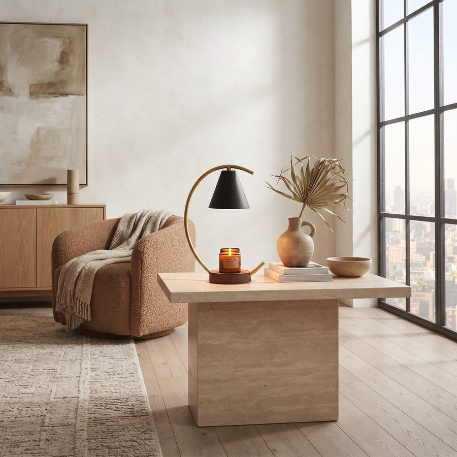 A modern minimalist living room scene featuring a light wood coffee table with a sculptural curved table lamp, a ceramic vase with dried palm leaves, and a candle on top. In the background, a textured tan armchair with a beige throw blanket sits next to a wooden sideboard under a framed abstract painting, all set in a bright room with a large multi-pane window.
