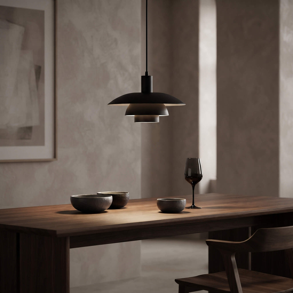 A modern black pendant light with a wide, layered disc shade hangs over a wooden dining table in a minimalist room with textured beige walls. The table is styled with dark bowls and a wine glass.