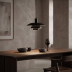 A modern black pendant light with a wide, layered disc shade hangs over a wooden dining table in a minimalist room with textured beige walls. The table is styled with dark bowls and a wine glass.