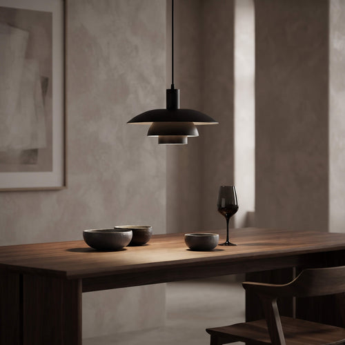 A modern black pendant light with a wide, layered disc shade hangs over a wooden dining table in a minimalist room with textured beige walls. The table is styled with dark bowls and a wine glass.