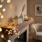 Warm white string fairy lights draped over a wooden mantel in a cozy living room setting, styled with books, a white vase, a dried grass stem, and a lit candle.