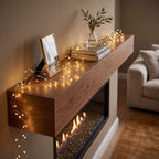 A warm wooden mantel shelf in a living room setting, decorated with glowing fairy lights, books, a small vase with greenery, and a framed picture above an electric fireplace. A beige sofa is visible in the background.
